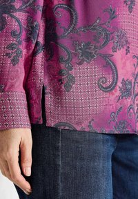 Roze blouse met een patroon, gekenmerkt door ingewikkelde zwarte bloemen- en paisleypatronen, met een zijopening en een contrasterende gepatterde manchette, gecombineerd met donkere denim.