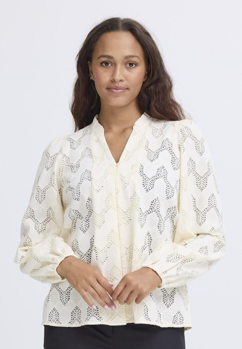 Cream blouse featuring a geometric lace pattern, V-neck, and gathered long sleeves. Textured fabric with subtle openness and button details.