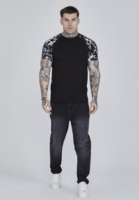 Mustat t-paidat, joissa on kukkakuosiset hihat, yhdistettynä tummiin farkkuihin ja valkoisiin lenkkareihin. Ominaisuuksiin kuuluu slim fit -design ja lyhyet hihat.