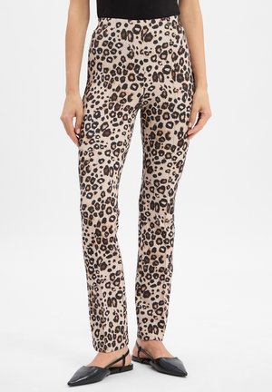 Pantalons en imprimé léopard beige avec des taches marron foncé et noires. Design ajusté avec une texture lisse. Portés avec des chaussures plates noires.