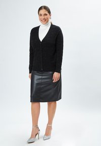 Cardigan nero texturizzato con scollatura a V e bottoni, abbinato a una gonna in pelle nera aderente e tacchi glitter argento. Finitura liscia e lucida.