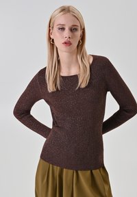 Femme blonde portant des boucles d'oreilles en or, un pull ajusté en ribbed marron et un pantalon plissé olive, posant les mains derrière le dos.