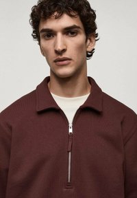 Burgundská mikina s vysokým límcem na zip a kontrastní krémovou vnitřní vrstvou, vyrobená z texturované tkaniny, s detailem stříbrného zipu.