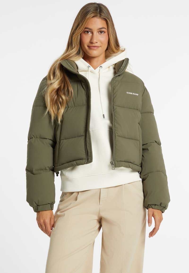 Alyvuogių žalios spalvos trumpa pūkinė striukė su užtrauktuku, elastingomis rankovėmis ir tekstūruotu audiniu. Pirmoje pusėje yra „Guess Jeans“ logotipas.
