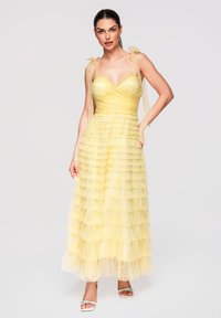 Femme en robe en tulle jaune à étages avec bretelles nouées aux épaules, debout, une main sur la hanche, portant des talons blancs à bouts ouverts.