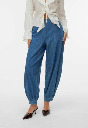 Frau trägt blaue, hoch taillierte, lockere Jeans mit elastischen Bündchen, kombiniert mit einer weißen Bluse mit Rüschen und dunklen spitzen Absatzschuhen.