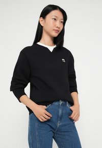 Sort sweatshirt med V-hals og lange ærmer, med en hvid inderside på kraven og et lille broderet logo på brystet. Båret sammen med blå jeans.