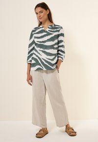Blouse rayée verte et blanche en tissu léger, avec un col en V et des manches trois-quarts, portée avec un pantalon large de couleur claire et des sandales.