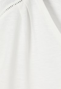 Tissu de vêtement blanc montrant une fine texture en maille, une couture diagonale et un petit détail de point ouvert près de l'encolure.