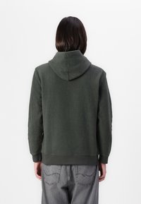 Hanorac din fleece verde închis cu glugă, material texturat, manșete elastice și tiv, purtat cu blugi gri deschis cu detalii cusute.