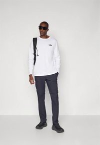 Balta garām piedurknēm krekls ar logotipu, tumšas kabatas bikses, melni apavi un saulesbrilles; rokās melna mugursoma; stāv pret gaišu fonu.