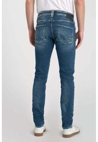 Jeans de denim azul de corte slim, con un acabado desgastado, cinco bolsillos y costuras en contraste. Combinados con una camisa blanca sencilla y zapatos casuales.