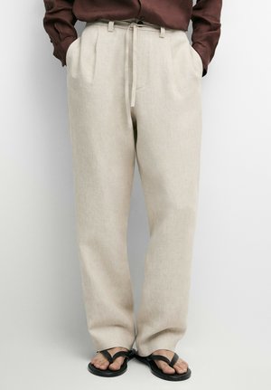 Person iført beige løstsiddende hørbukser med snøre i taljen, kombineret med mørkebrun skjorte og sorte åbne sandaler.