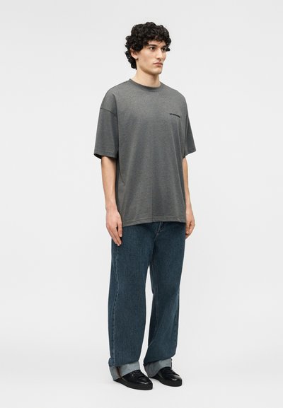 Tânăr cu păr creț, purtând un tricou gri larg, blugi largi de culoare albastru închis cu tiv răsucit și teniși negri, stând într-o poziție neutră pe un fundal alb.