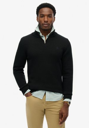 Pull noir en maille avec un col zippé, associé à une chemise rayée bleu clair et blanc. Porté sur un pantalon kaki. Design simple.