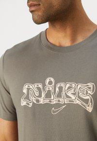 Szürke pamut póló, amelynek elülső oldalán egy nagy, stilizált fehér "Nike" logó található, alatta pedig egy kis Nike pipa, kerek nyakú dizájn.