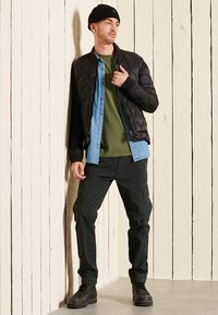 Zwarte gewatteerde bomberjack over een groen T-shirt en denim overhemd, gecombineerd met donkere cargobroeken en zwarte laarzen. Een beanie maakt de look compleet.