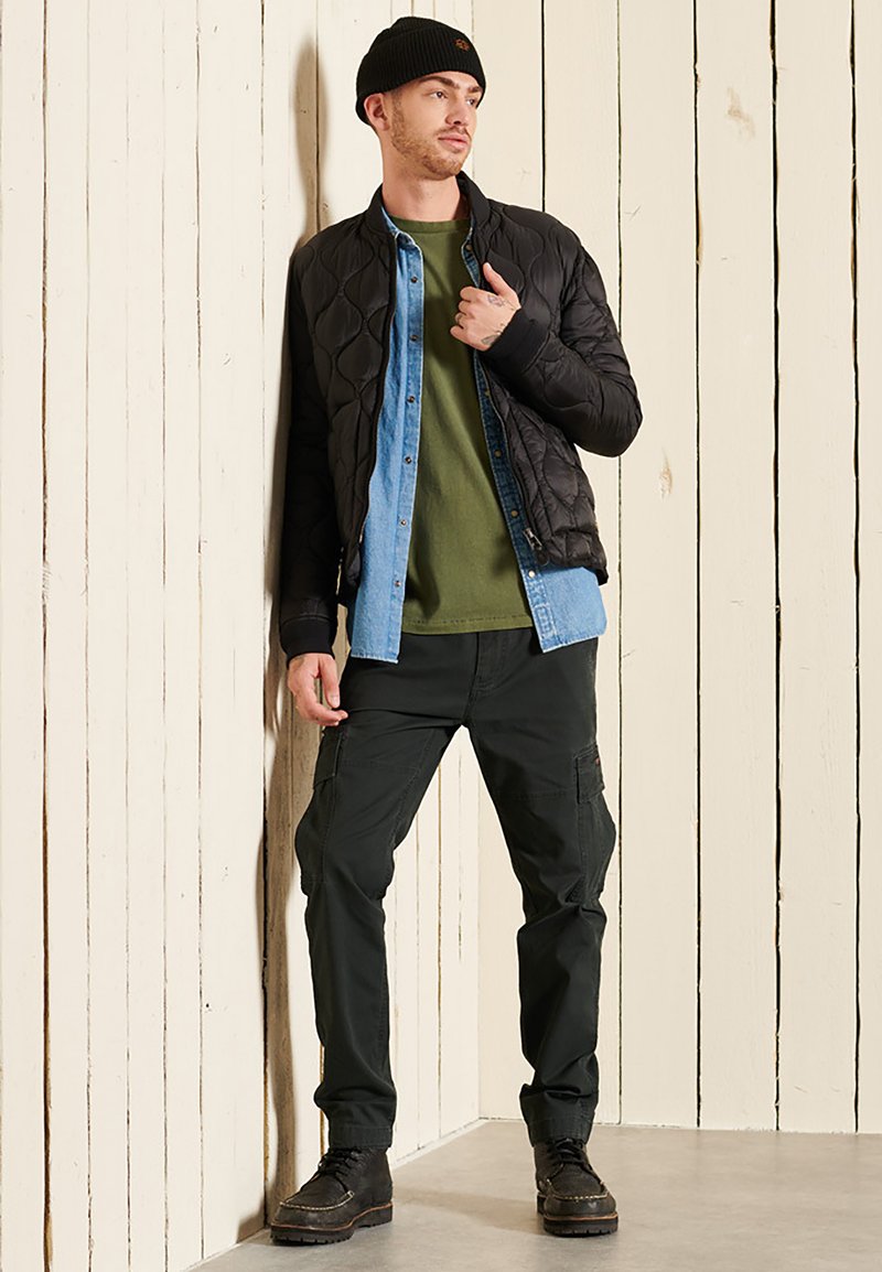 Zwarte gewatteerde bomberjack over een groen T-shirt en denim overhemd, gecombineerd met donkere cargobroeken en zwarte laarzen. Een beanie maakt de look compleet.