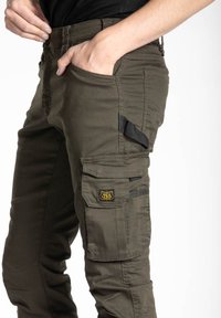 Pantalons cargo en vert olive, avec une fermeture à bouton, de multiples poches, un tissu texturé, et un logo en patch sur la poche latérale.