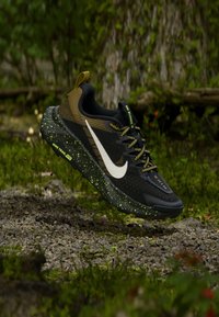 Παπούτσια trail running Nike, με μαύρο πάνω μέρος και ελαιώδεις λεπτομέρειες, άσπρο λογότυπο και σπειροειδές σόλο, σχεδιασμένα για εξωτερική απόδοση.