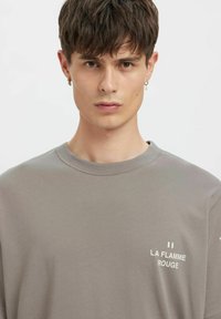 Grå bomulls-T-skjorte med rund hals og korte ermer. Har hvit tekst "LA FLAMME ROUGE" på fronten. Jevn tekstur, avslappet design.