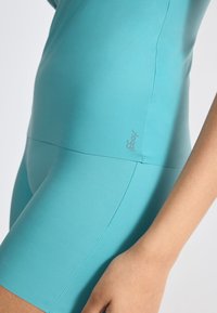 A close-up of a fitted, short turquoise activewear top, featuring smooth, stretchy fabric and a subtle logo near the hem.