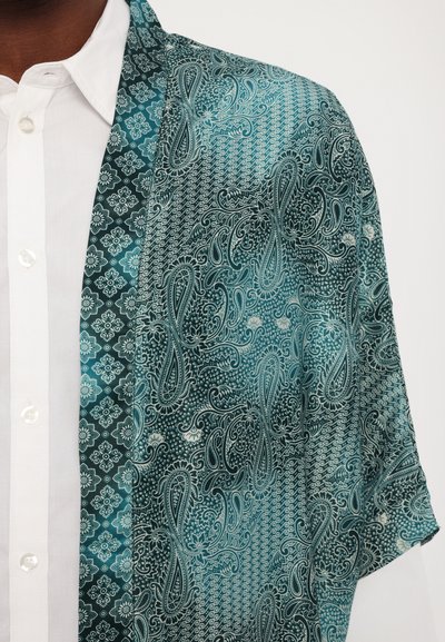 Svileni kimono v teal in črni barvi z zakompliciranimi paisley in geometrijskimi vzorci. Ima mehko teksturo in tekoč dizajn, delno nanesen nad belo srajco.