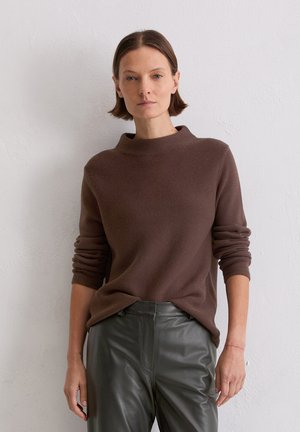 Brązowy, teksturowany sweter z wysokim dekoltem, noszony na ciemnozielonych skórzanych spodniach. Długie rękawy są lekko podwinięte. Prosty, nowoczesny design.