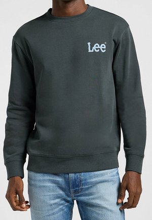 Mężczyzna noszący ciemnoszarą bluzę Lee z jasnoniebieskim logo na piersi i jasnoniebieskie jeansy, stojący na jednolitym tle.