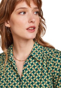 Blusa verde con motivo geometrico, arricchita da dettagli gialli e rossi. Dotata di colletto e bottoni. Collana in catena dorata mostrata.