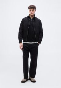 Νέος άνδρας που στέκεται φορώντας μαύρο μπουφάν με φερμουάρ, μαύρο πουκάμισο, μαύρο παντελόνι και γκρι παπούτσια χωρίς κορδόνια σε απλό λευκό φόντο.