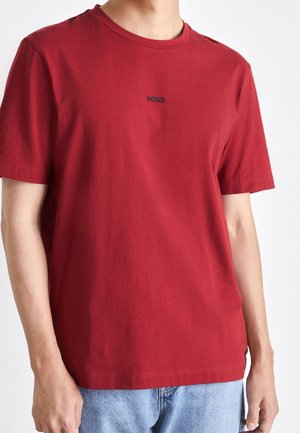 Mann trägt ein rotes T-Shirt mit kurzen Ärmeln und "BOSS" Logo in der Mitte der Brust sowie hellblaue Jeans vor weißem Hintergrund.