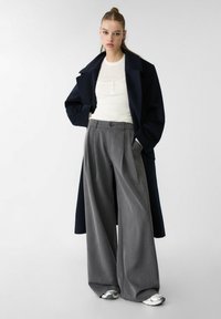 Marineblauer Überrock, weißes, tailliertes Oberteil und graue, weit geschnittene Hose. Akzente umfassen einen Knopfverschluss und silberne metallische Schuhe. Glatte Texturen.