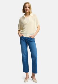 Pull léger en maille crème avec col, manches courtes et coupe ample, associé à un jean droit bleu et des mules beiges.