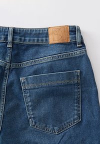 Jean en denim bleu avec une étiquette en cuir beige à la taille, des coutures contrastantes et une seule poche arrière. Texture douce et design classique.