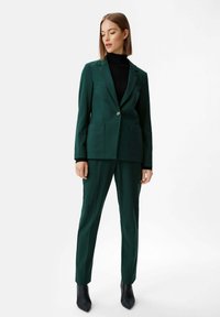 Green tailored blazer with two front pockets, matching tapered trousers, and a black turtleneck. Pointed black ankle boots complete the look.