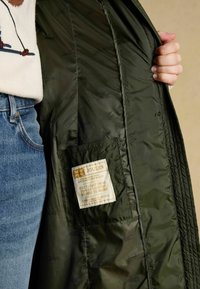 Prešívaná zelená bunda s hladkou textúrou a vnútornou vreckom s etiketou. V spojení so svetlomodrými rifľami.