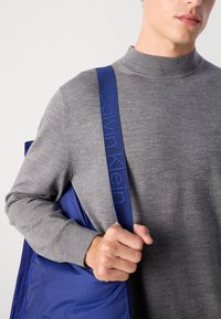 Grå strikket genser med høy krage, sammen med en blå nylonveske med en teksturert stropp preget med "Calvin Klein."