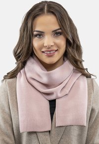 Eșarfă tricotată de culoare roz deschis, cu o textură moale și margini cu model, purtată peste un palton bej, căzând elegant în jurul gâtului.