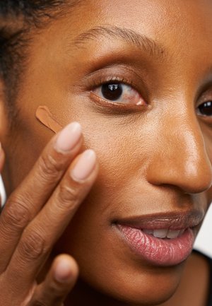 Fond de teint liquide appliqué sur la peau du visage ; texture lisse ; couleur beige chaude ; main du modèle avec vernis à ongles rose clair ; mise au point sur la zone de la joue.