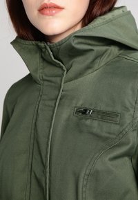 Veste vert olive avec un col montant et une poche zippée. Le matériau semble lisse avec des coutures visibles et une coupe structurée.