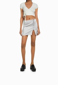 Vit ribbad kort wrap-topp med knytdetalj, parat med en silverfärgad satäng-minikjol med en sidaöppning och knappdekorationer. Svarta platåklackar.