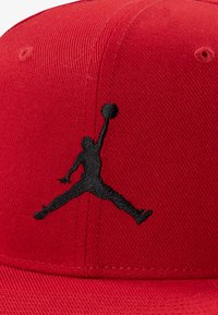 Red cap made of textured fabric, featuring a black embroidered Jumpman logo. The shape is structured with a flat brim and stitched panels.