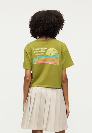 Άτομο με σγουρά μαλλιά φοράει ελαιί πράσινο cropped t-shirt με πολύχρωμο γραφικό κύμα και λευκή πλισέ φούστα, στραμμένο προς τα πίσω.