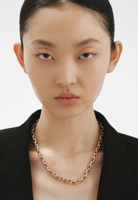 Collar de cadena de oro con eslabones entrelazados, llevado sobre un blazer negro, revelando piel suave y un fondo neutro.