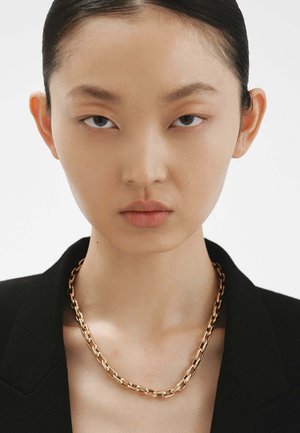 Collar de cadena de oro con eslabones entrelazados, llevado sobre un blazer negro, revelando piel suave y un fondo neutro.