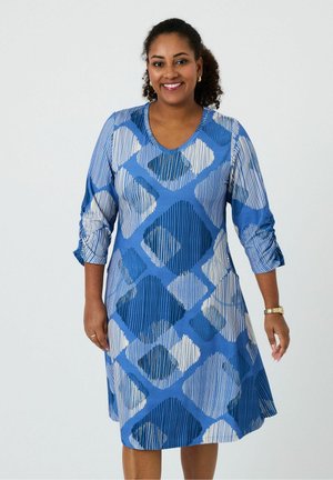 Smiling woman wearing blue and white geometric patterned dress with three-quarter sleeves, standing against a plain light background.