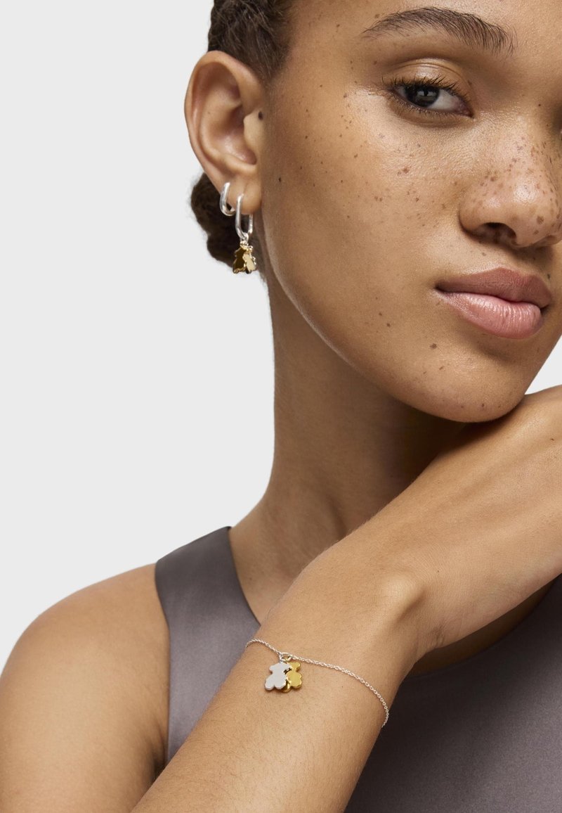 Pulsera de cadena de plata con dos charms: uno de plata y otro de oro, junto con pendientes de aro con acentos dorados que tienen un charm. Textura de piel suave.