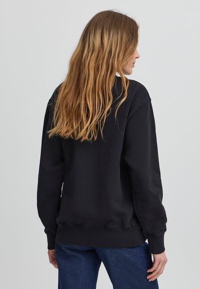 Fekete pulóver laza szabással, bordázott mandzsettákkal és aljjal. Simább textúrája és klasszikus crew nyaka van. Hátsó nézet hosszú hajjal.