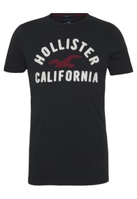 Camiseta de algodón negra con texto bordado en blanco y burdeos "HOLLISTER CALIFORNIA" y un gráfico de un águila. Manga corta, cuello redondo.